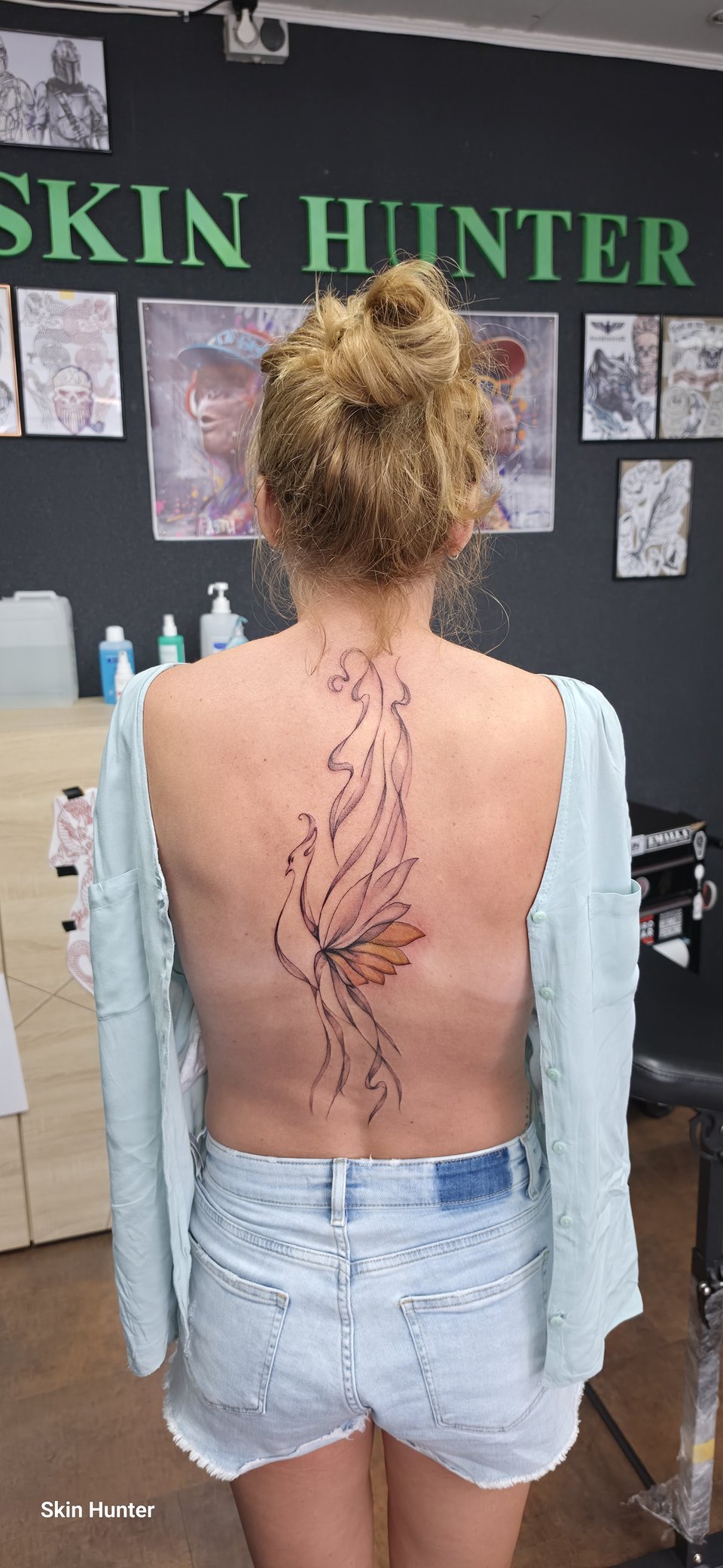 Florales Rücken-Tattoo in feinen Linien mit orangen Akzenten vor dem Skin-Hunter-Schriftzug